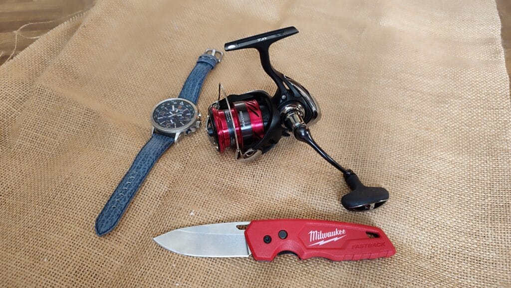 Photo réelle moulinet Daiwa Ninja 2500 LT : comparaison de taille avec une montre homme et un couteau Milwaukee Fastback sur fond brut.